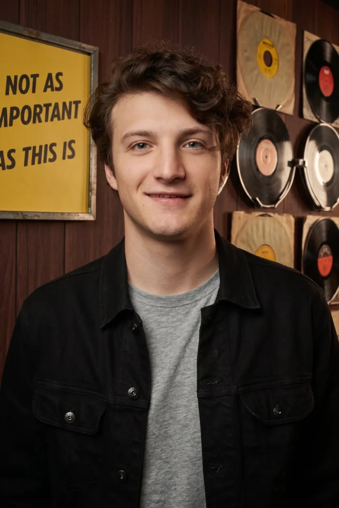 A smiling young man wearing a black denim jacket standing in front of a wall with vintage vinyl records and a yellow sign.