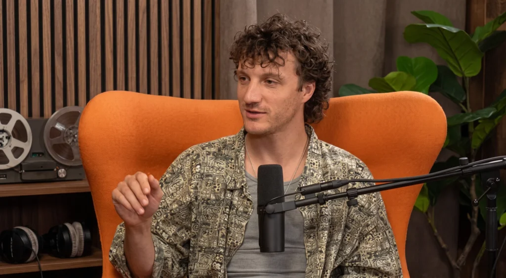 A young man with curly hair wearing a patterned shirt, speaking into a microphone while sitting in an orange chair during a podcast.