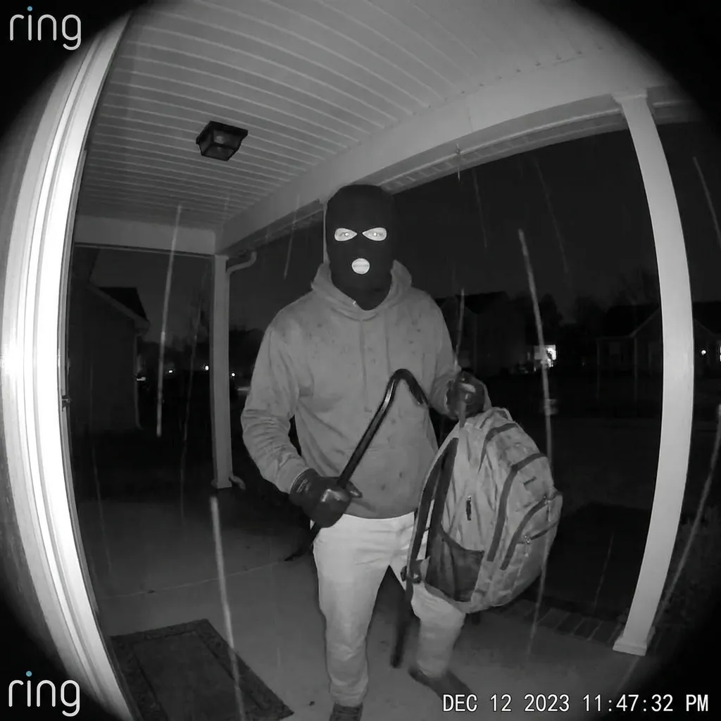 Black and white Ring doorbell surveillance footage of a masked man standing on a porch holding a weapon and a black backpack.