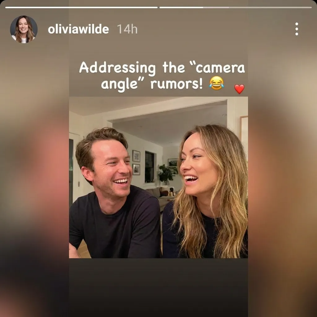 Screenshot of Olivia Wilde's Instagram story showing her laughing with a male companion indoors, with text reading 'Addressing the camera angle rumors!' accompanied by a laughing emoji and red heart.