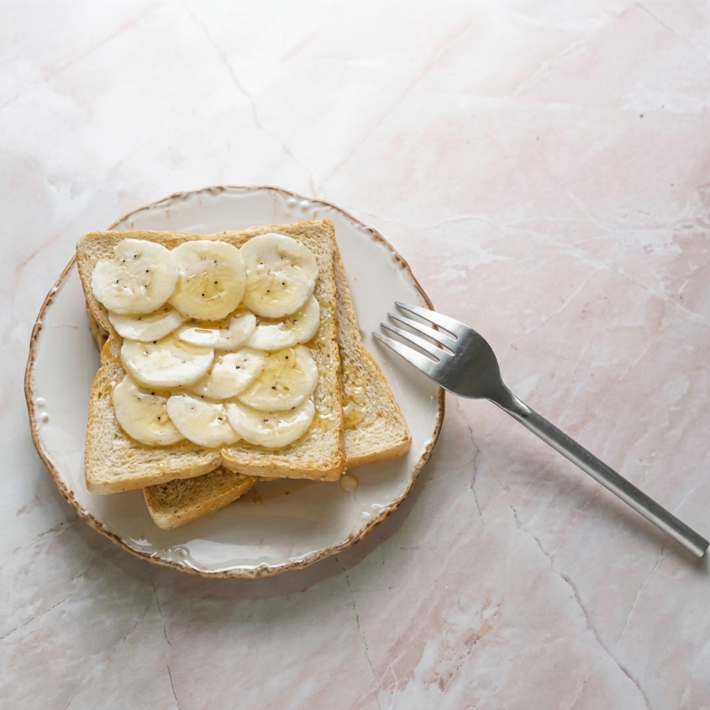 Top-down view of an open-faced sandwich with sliced bananas on toast, served on a rustic white plate next to a silver fork on a pink marble countertop.