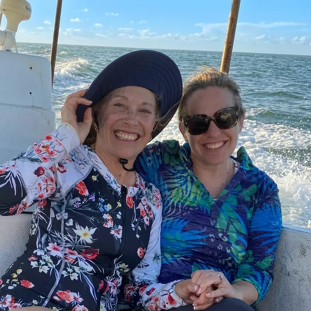 Smiling senior mother and adult daughter enjoying a sunny boat ride on the ocean, wearing a sun hat and sunglasses.