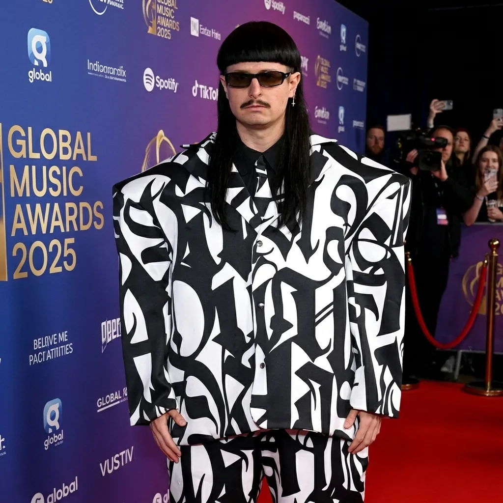 : Musician Oliver Tree posing on the red carpet at the Global Music Awards wearing a custom oversized black and white patterned suit, dark sunglasses, and his signature bowl cut hairstyle.
