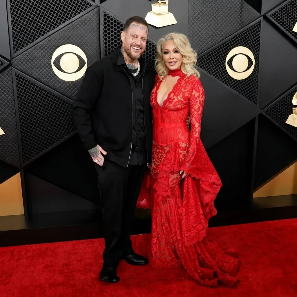 Jelly Roll and Bunnie XO standing on a red carpet in front of a black and gold Grammy Awards backdrop. Jelly Roll is wearing an all-black outfit with a jacket, and Bunnie XO is wearing a bright red, intricate lace gown with a corset-style bodice.