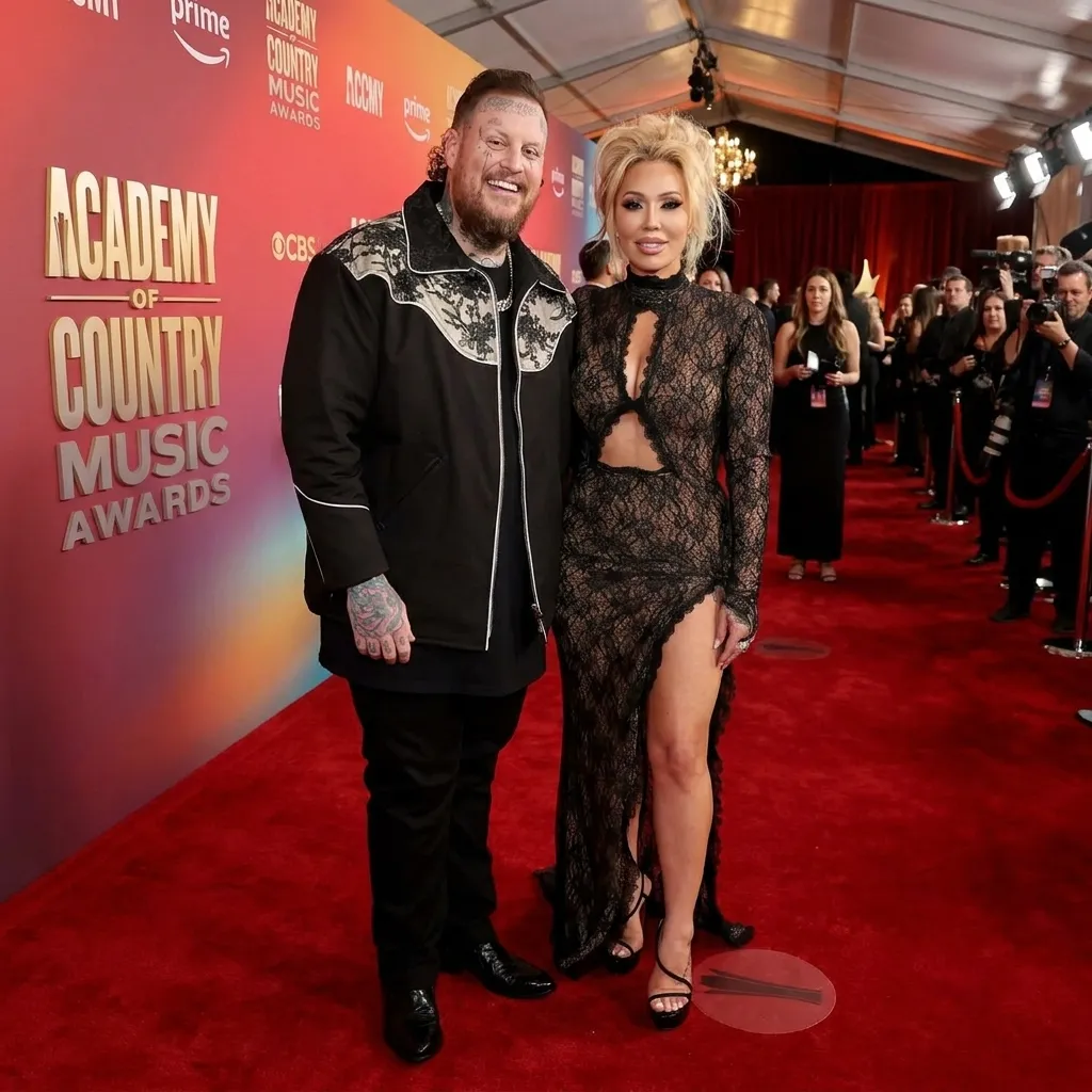 Jelly Roll and Bunnie XO posing on the red carpet at the Academy of Country Music Awards. Jelly Roll is wearing a black western-style jacket with white embroidery, and Bunnie XO is wearing a black lace gown with cutouts and a high leg slit.