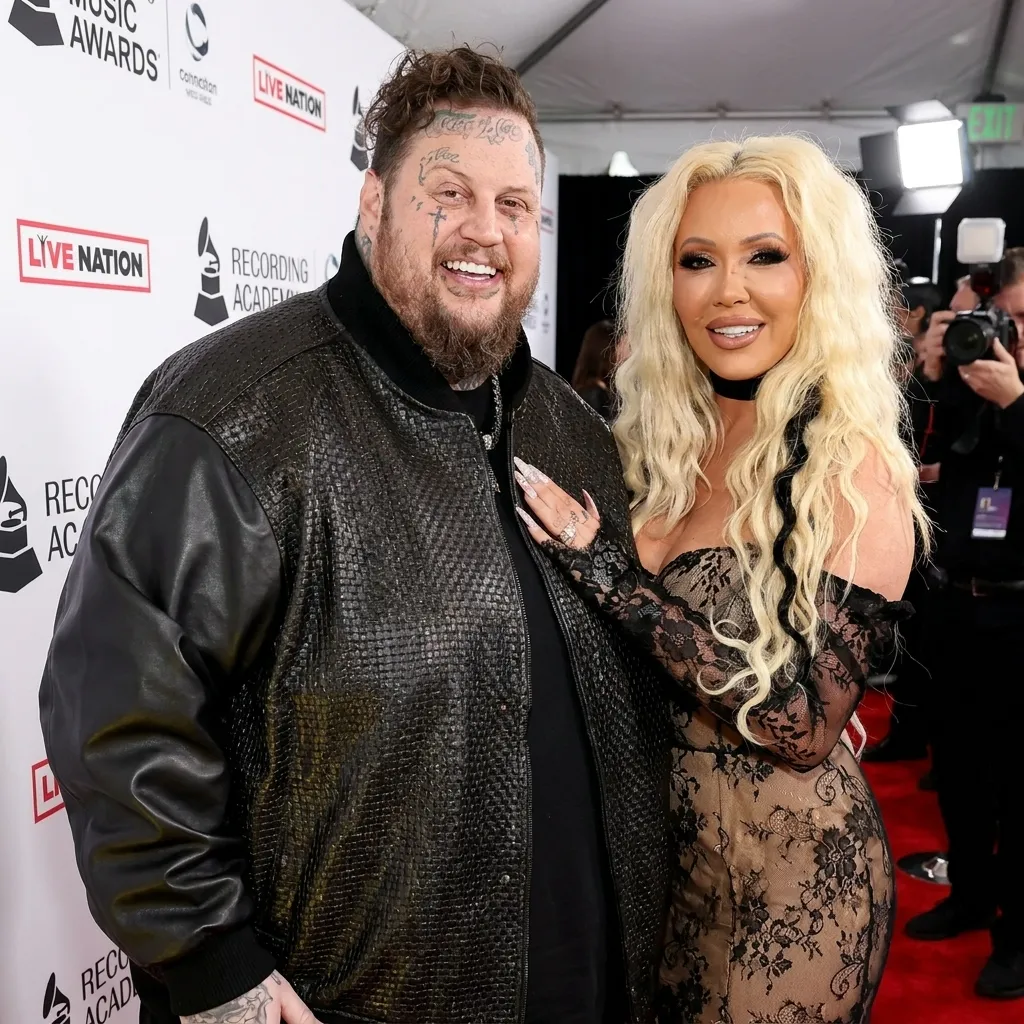 Jelly Roll and Bunnie XO smiling together on a red carpet. Jelly Roll is wearing a textured black bomber jacket, and Bunnie XO is wearing a black lace off-the-shoulder dress. The background features "Live Nation" and "Recording Academy" logos.