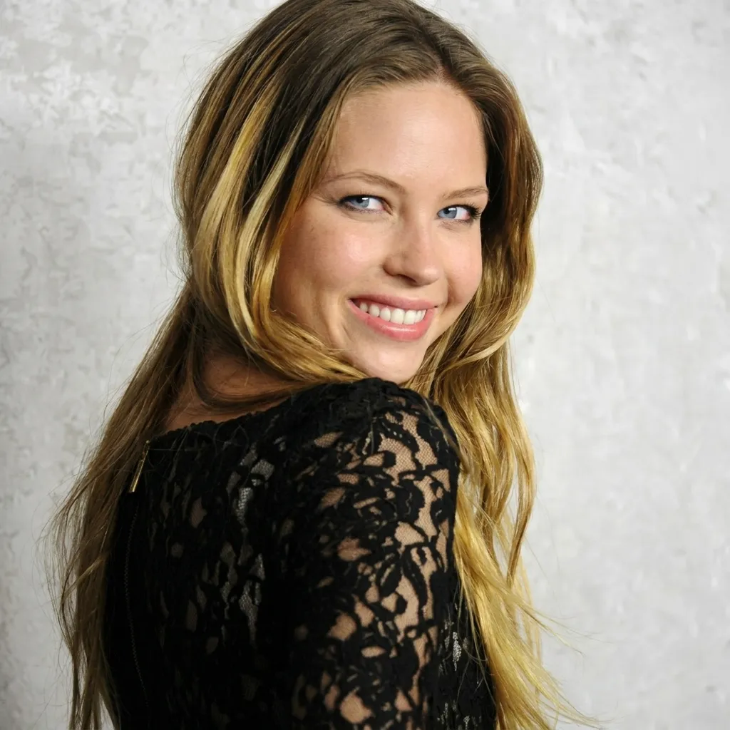 Smiling young blonde woman with blue eyes wearing a black lace top, looking over her shoulder against a white textured background.