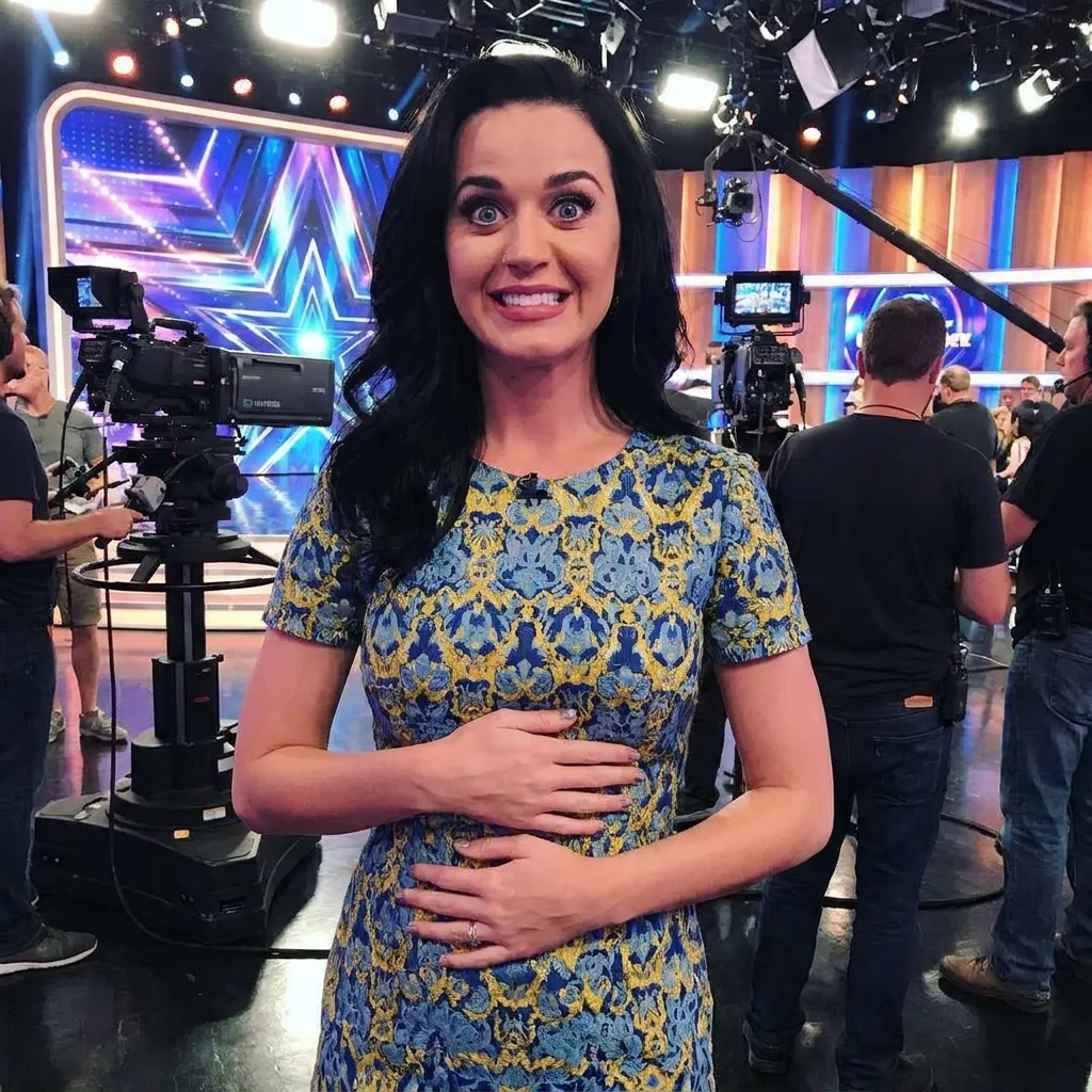 Katy Perry making a wide-eyed, funny expression while holding her blue and yellow patterned dress on a brightly lit television studio set