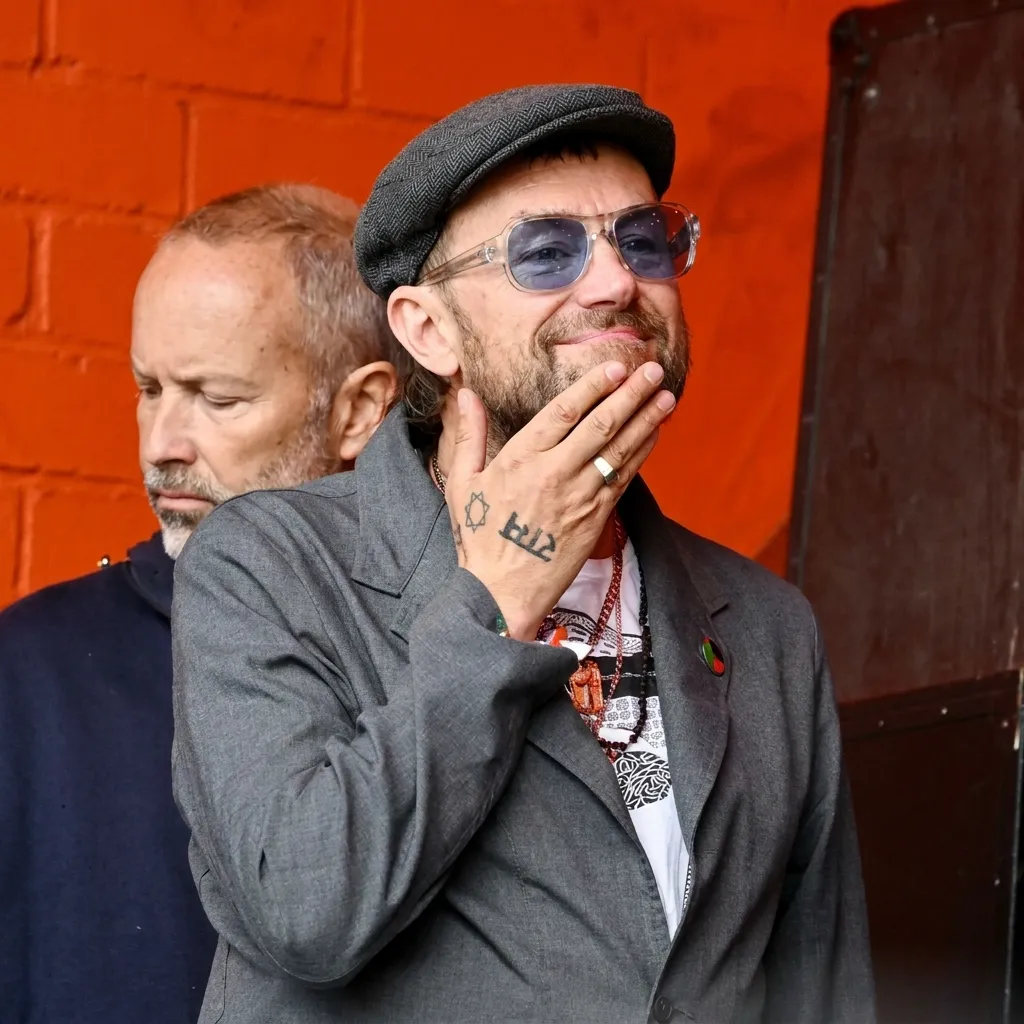 Damon Albarn smiling and touching his chin, wearing a grey jacket, flat cap, and blue-tinted sunglasses in front of an orange wall