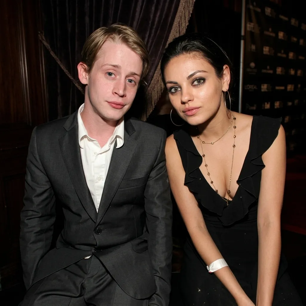 Macaulay Culkin wearing a dark suit and Mila Kunis wearing a black dress posing together at an indoor event.