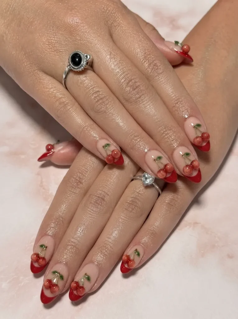 Two hands with almond-shaped nails featuring red French tips embellished with 3D glossy cherry art and green stems, wearing silver statement rings.