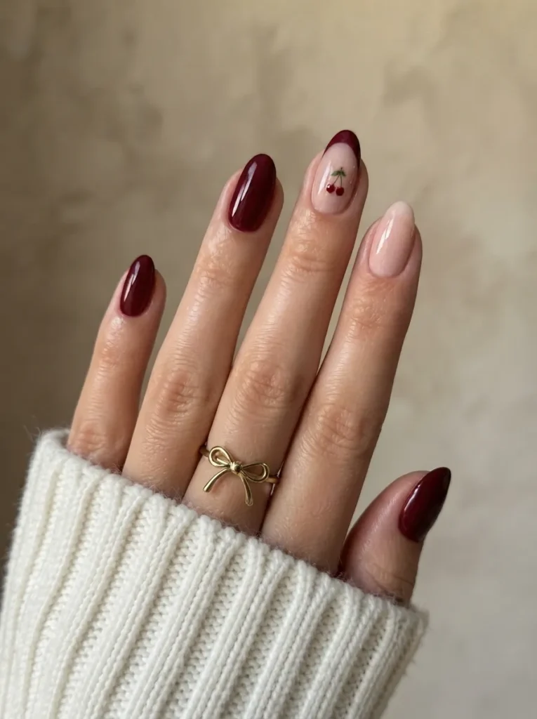 Hand wearing a white ribbed sweater and a gold bow ring, displaying a manicure with solid deep burgundy nails, a solid nude nail, and a burgundy French tip nail with a delicate cherry accent.