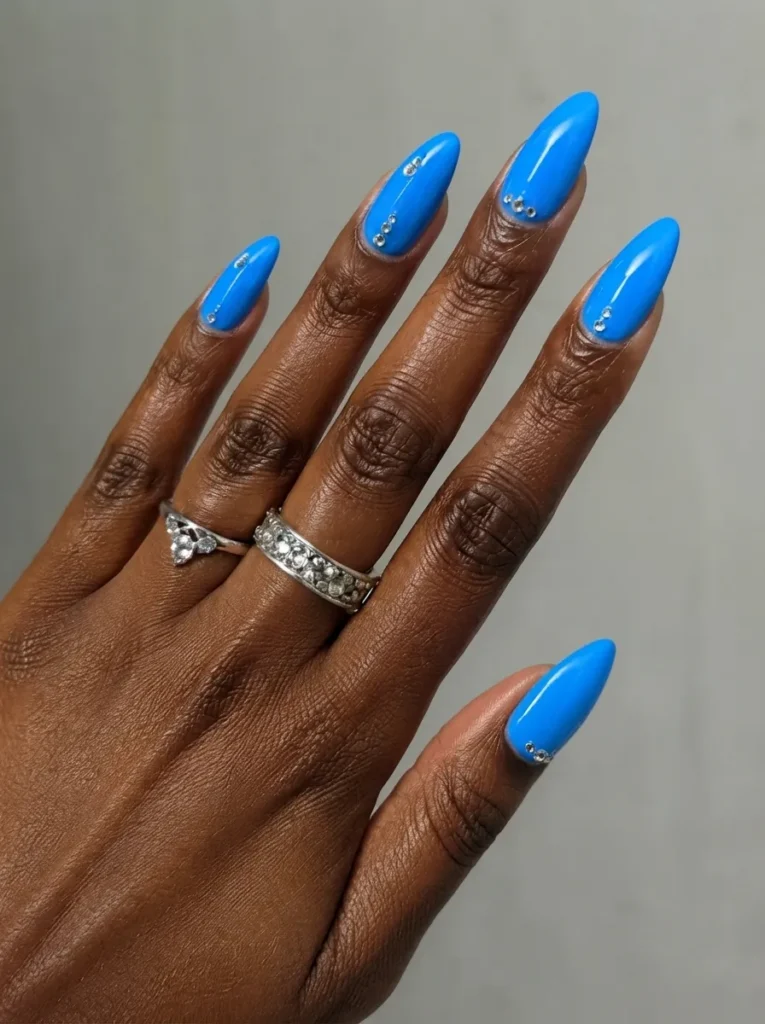 Almond-shaped nails painted in a solid vibrant blue with delicate rhinestone accents near the cuticles