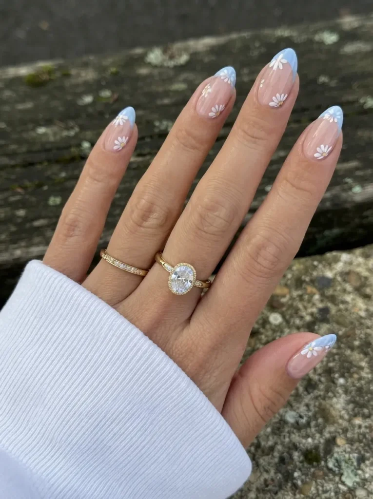 Almond-shaped nails with light blue French tips and painted white daisies, styled with gold rings.