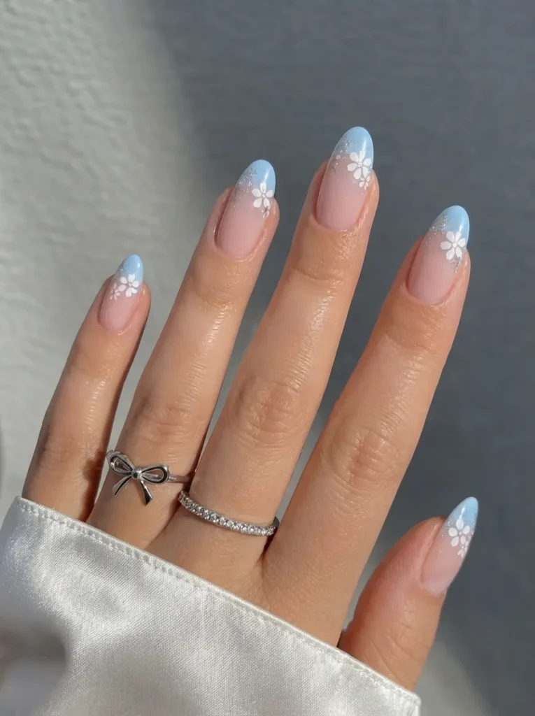 Almond-shaped nails with light blue tips, white flowers, and subtle silver glitter, worn with silver rings.