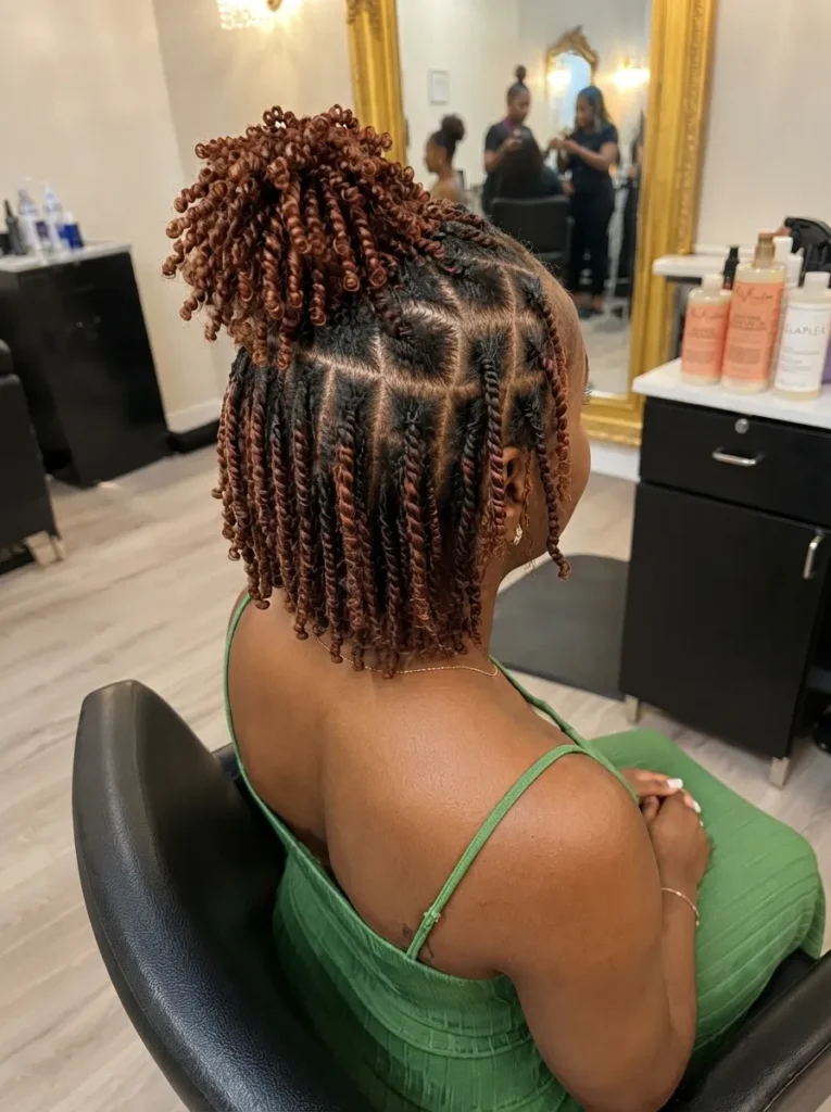 A woman in a green top sitting in a salon chair, viewed from behind to showcase her copper-brown kinky twists styled in a half-up ponytail with neatly parted box sections.
