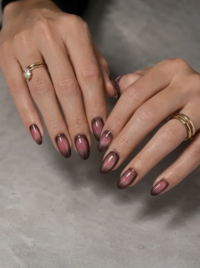 Hands showcasing dusty plum cat-eye almond-shaped nails accessorized with gold rings
