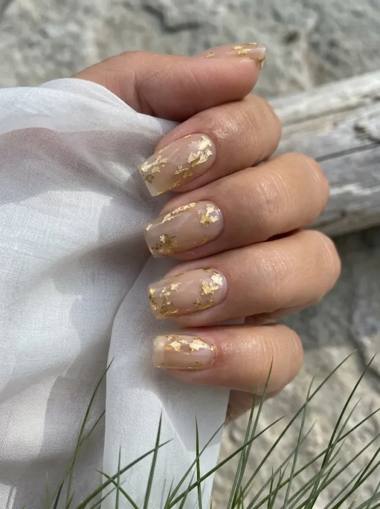 Hand holding a white fabric showcasing glossy nude coffin nails encapsulated with delicate gold foil.