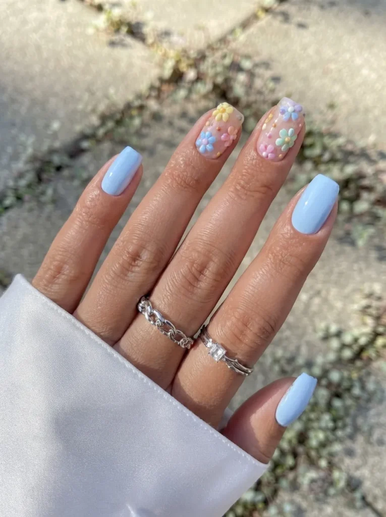 Light blue square nails featuring 3D pastel floral nail art designs, accessorized with silver rings.