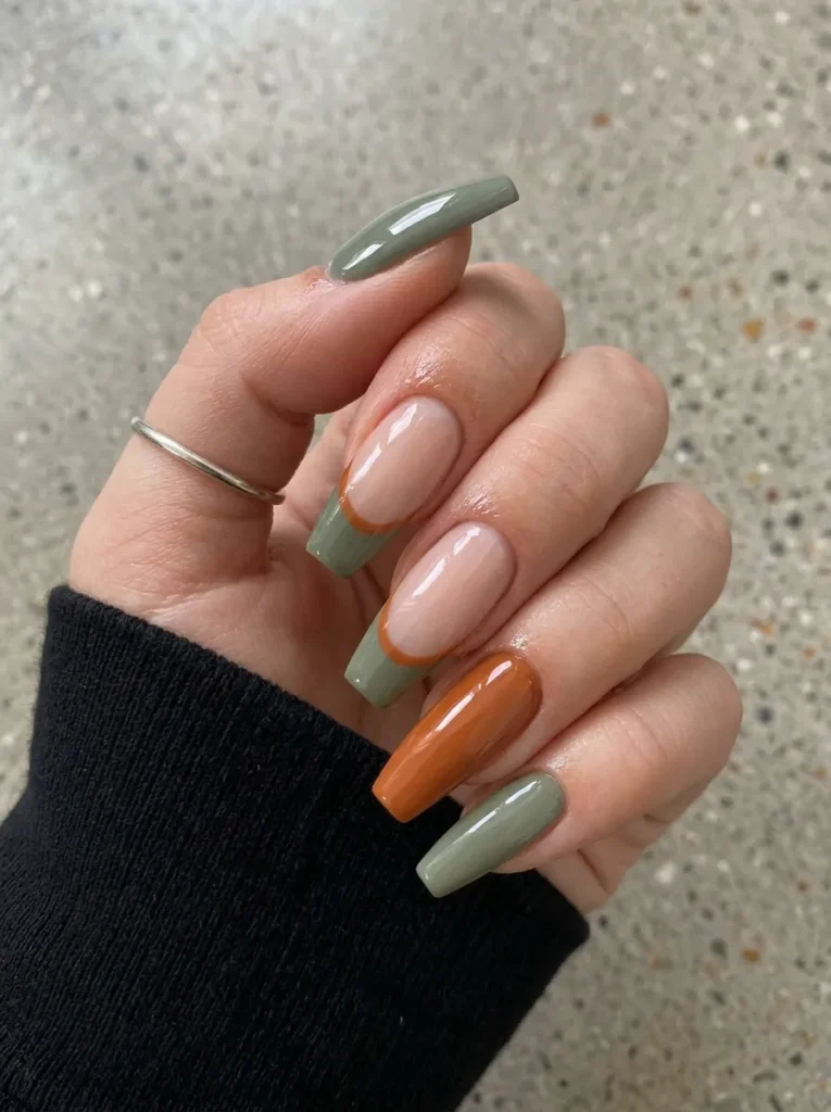 Hand in a black sleeve wearing long coffin nails combining solid sage green, burnt orange, and two-tone French tips