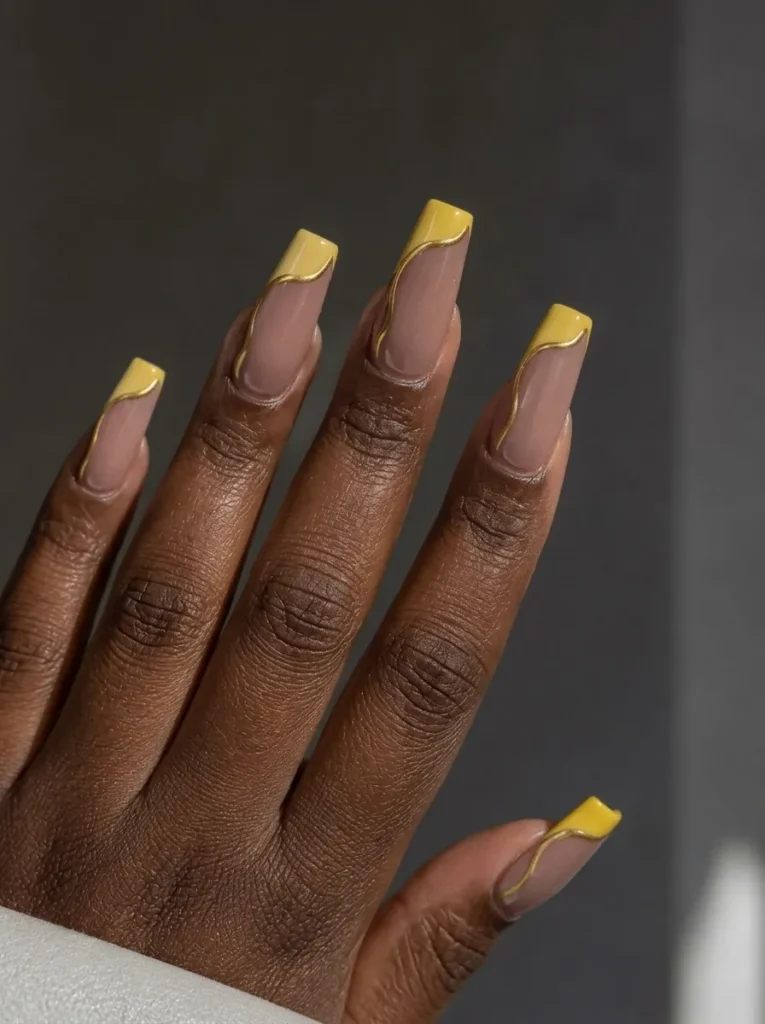 Long square nails with a nude base, yellow tips, and elegant gold foil swirl accents