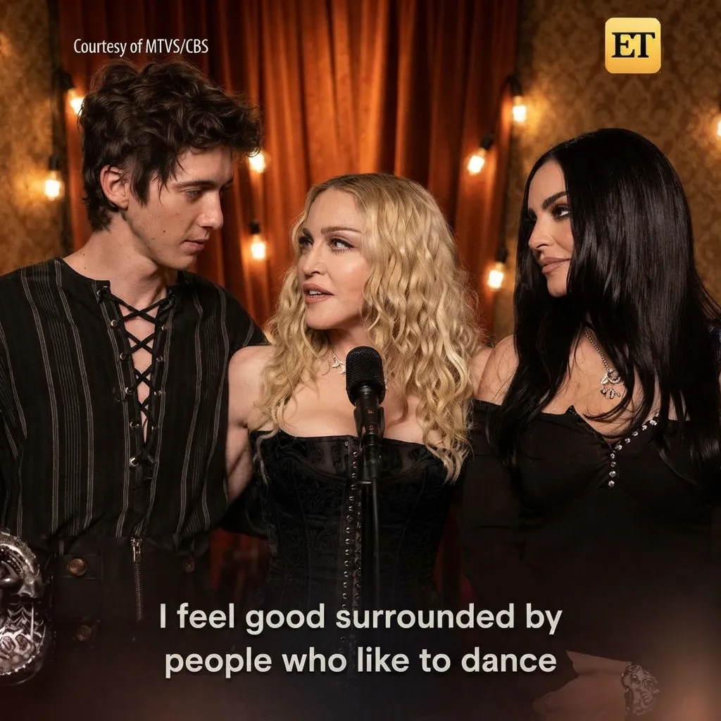 Madonna speaking into a microphone with two guests against a vintage backdrop with warm string lights.