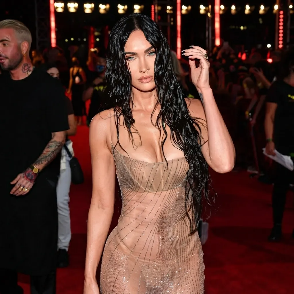 Megan Fox posing on a red carpet with wet-look dark hair and a sheer, sparkling nude-toned dress.