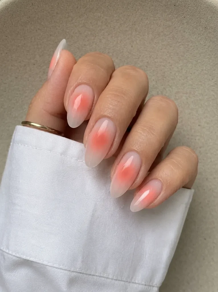 Hand showcasing milky white almond nails with vibrant coral blush aura centers and a gold ring