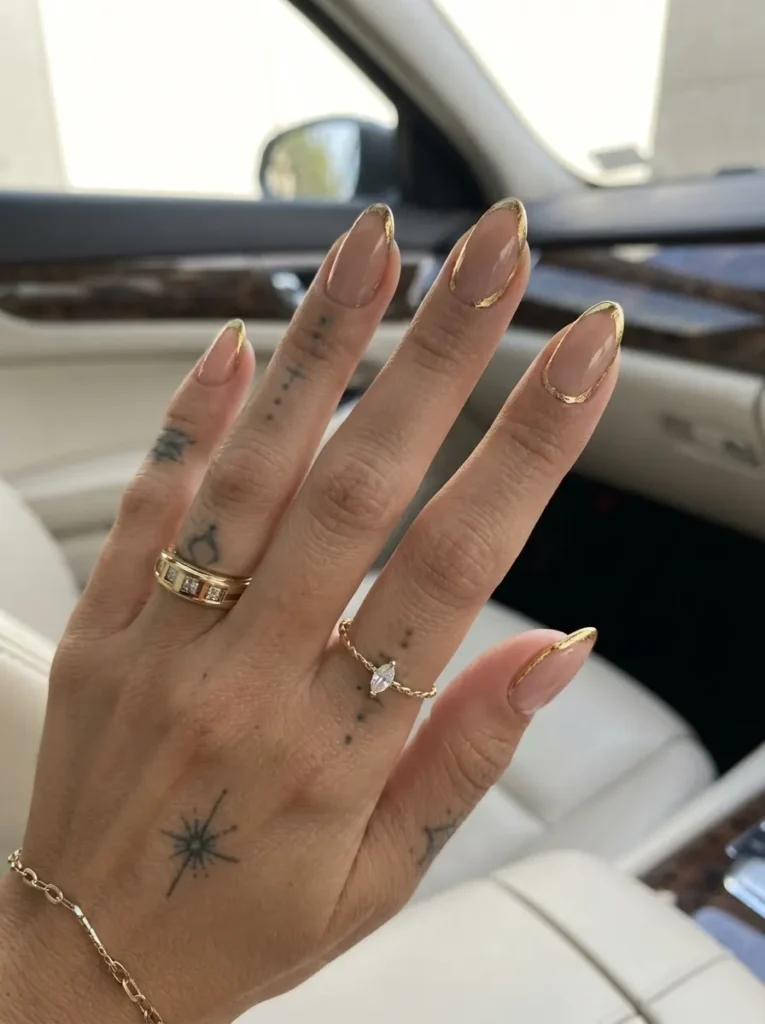 Hand with minimal nude almond-shaped nails featuring delicate metallic gold French tips, multiple finger tattoos, and gold rings