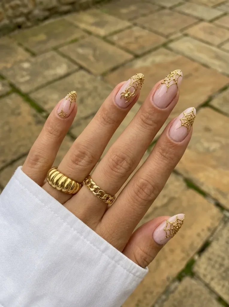 Hand adorned with nude almond nails featuring glowing gold spiderwebs and tiny spider accents.