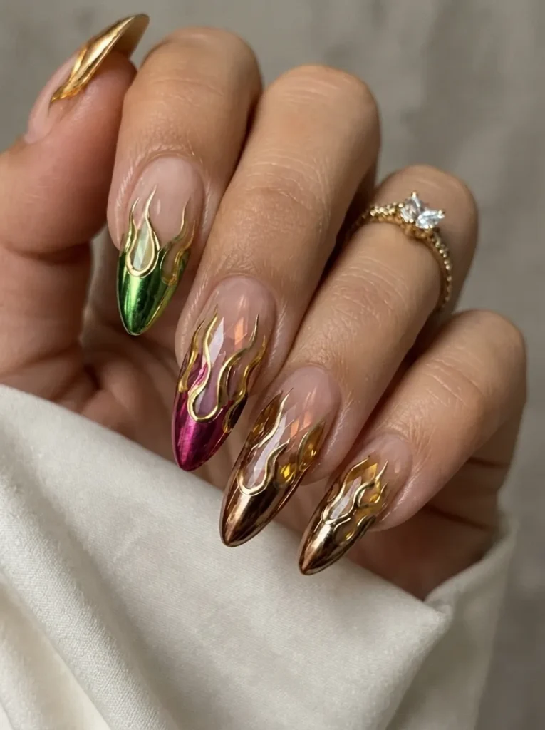 Hand showing almond-shaped nails with a sheer nude base and metallic gold 3D flame patterns outlining jewel-toned metallic tips.
