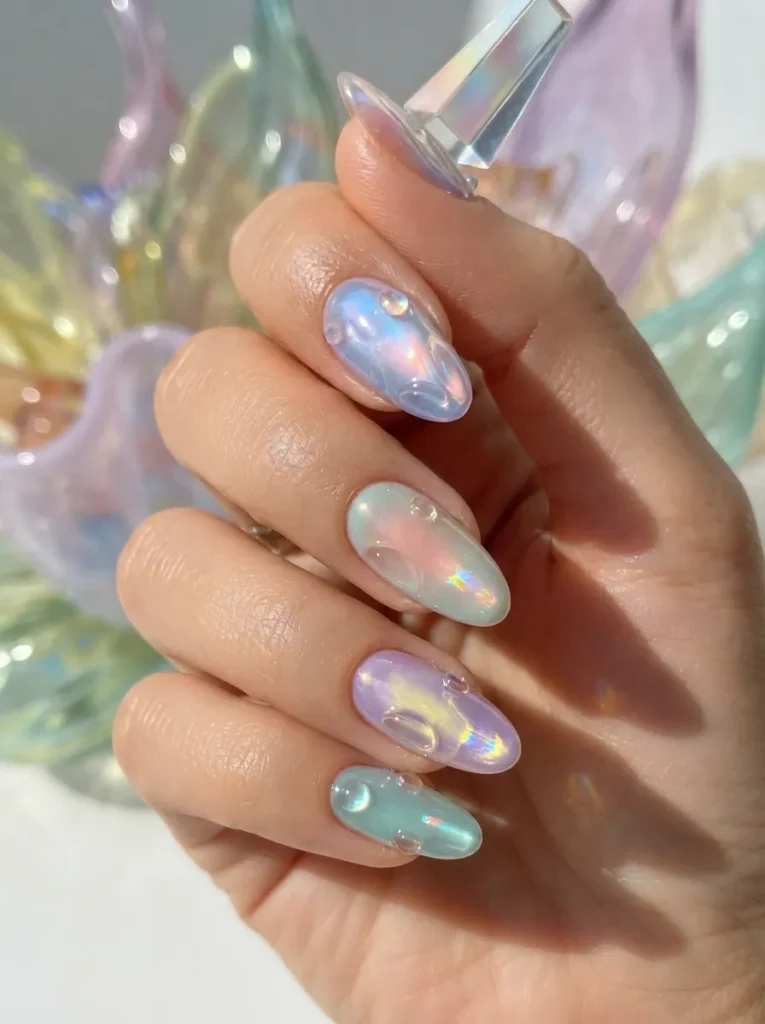 Hand displaying pastel iridescent aura nails with 3D water droplet effects against a colorful abstract glass background.