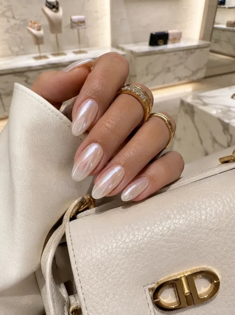 Hand with almond-shaped nails featuring a glossy pearl-glow chrome finish, wearing gold rings and holding a white leather designer handbag.