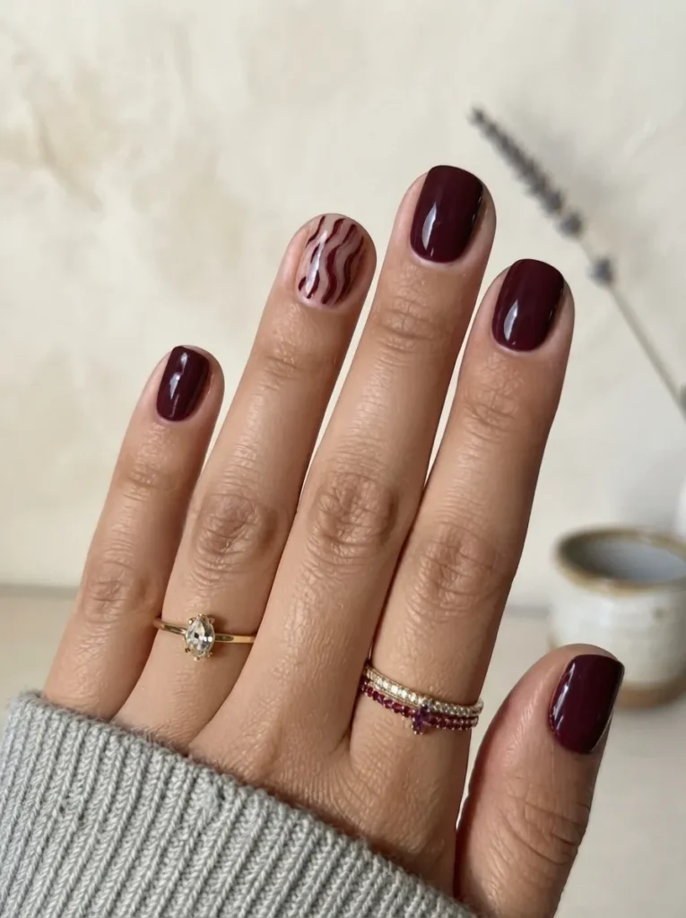 Short round burgundy nails with a nude and burgundy zebra stripe accent nail, adorned with gold rings.