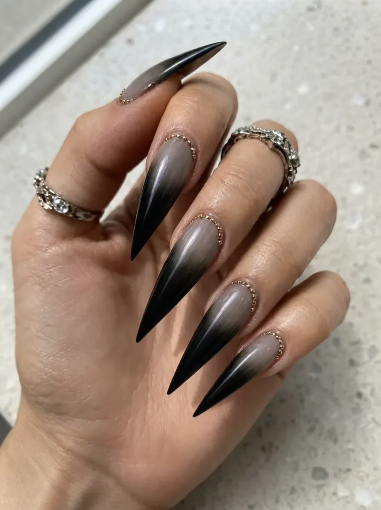 Long stiletto nails with a smoky black and grey ombre gradient, detailed with delicate gold micro-beads along the cuticles