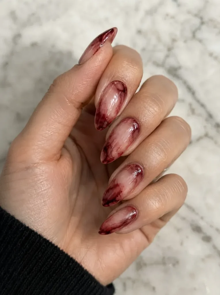 Almond-shaped nails featuring a moody, translucent watercolor marbling effect in deep crimson red and black over a nude base.