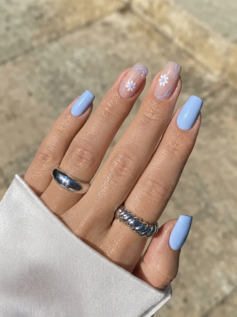 Square-shaped nails featuring solid light blue polish and delicate floral accent nail art, accessorized with silver rings.