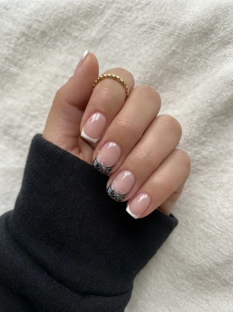 Short square nails featuring classic white French tips on some fingers, while others display delicate black spiderweb art along the tips.