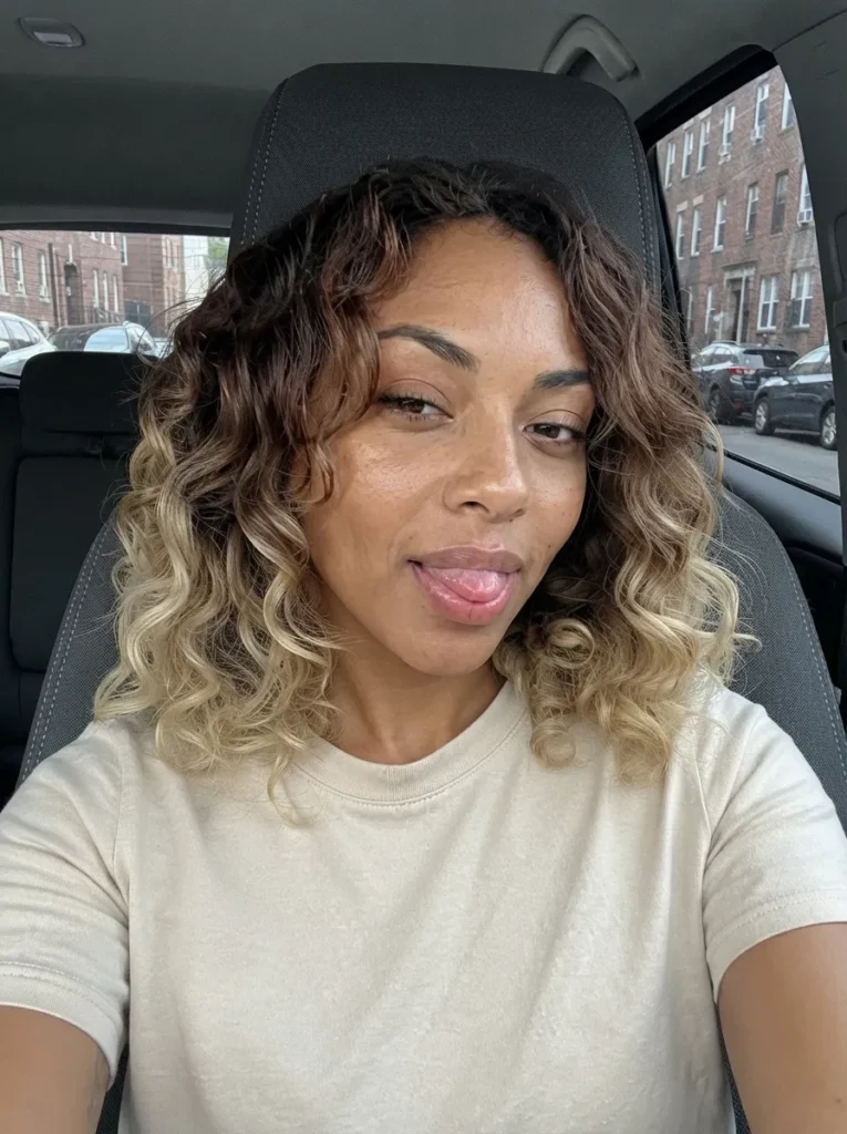 Selfie of a Black woman with brown-to-blonde ombre curly hair sticking her tongue out while sitting in a car.
