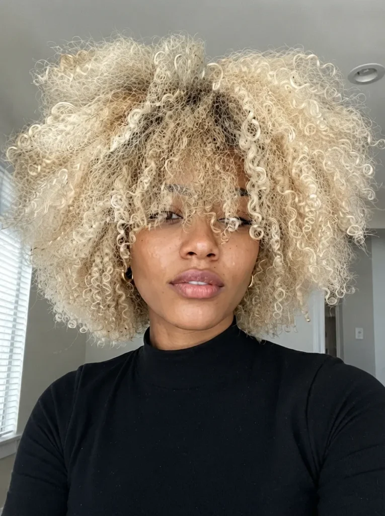 Portrait of a Black woman with a massive, voluminous light blonde curly afro and bangs, wearing a black high-neck top.