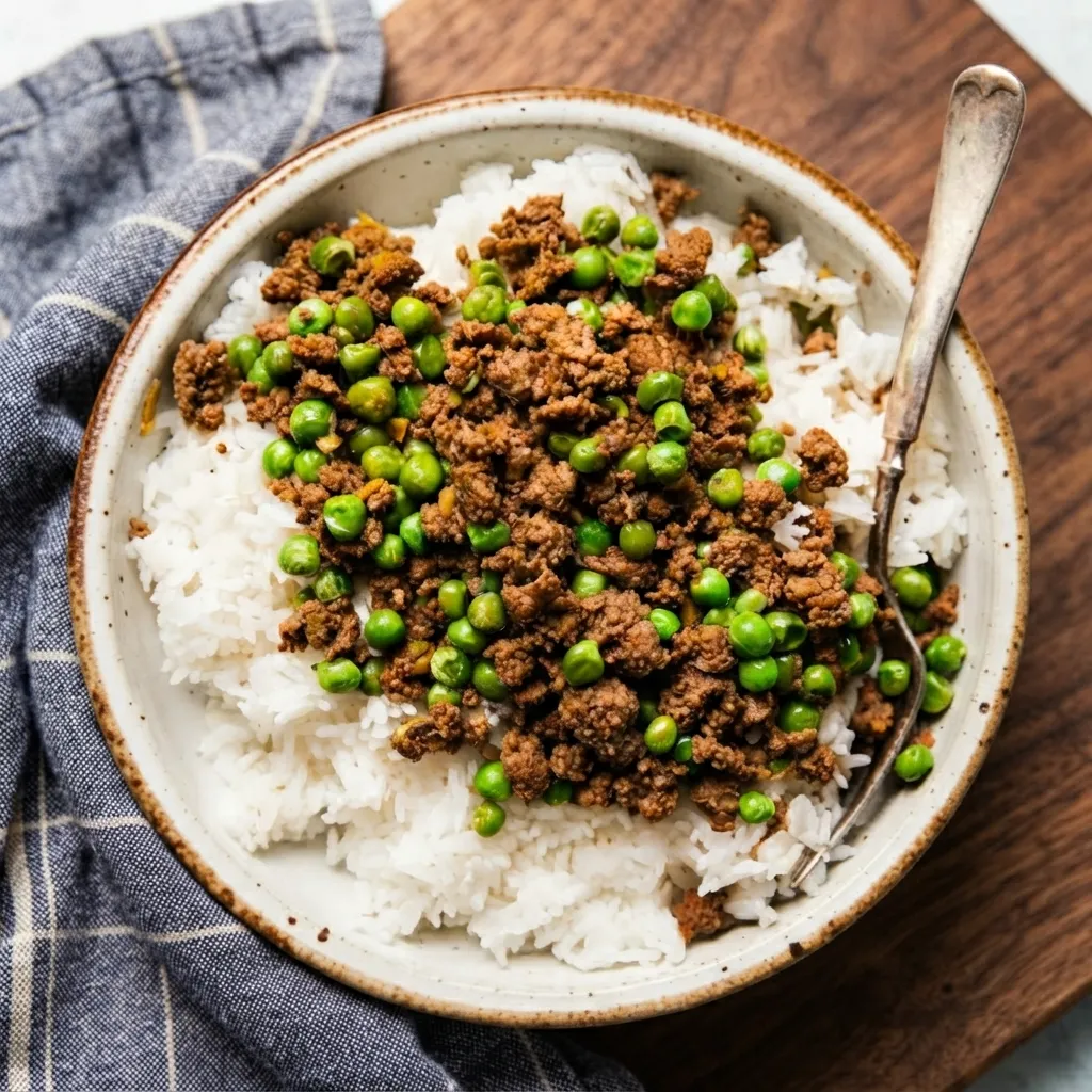 A bowl of fluffy white rice topped with seasoned ground beef and green peas, sitting on a wooden board next to a blue patterned napkin and a serving spoon.