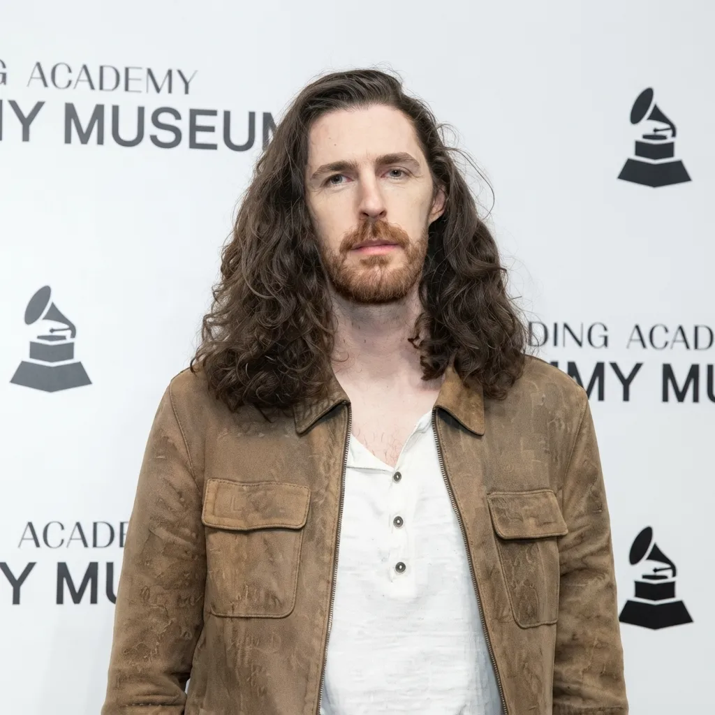 Hozier with long dark hair and a beard, wearing a white buttoned shirt and brown suede jacket in front of a Recording Academy / Grammy Museum backdrop.