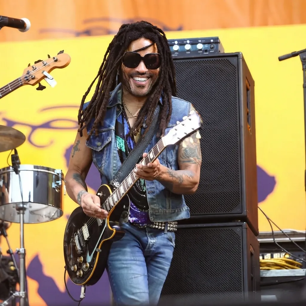 Lenny Kravitz smiling and playing an electric guitar on stage, wearing sunglasses, a denim vest, and dreadlocks.